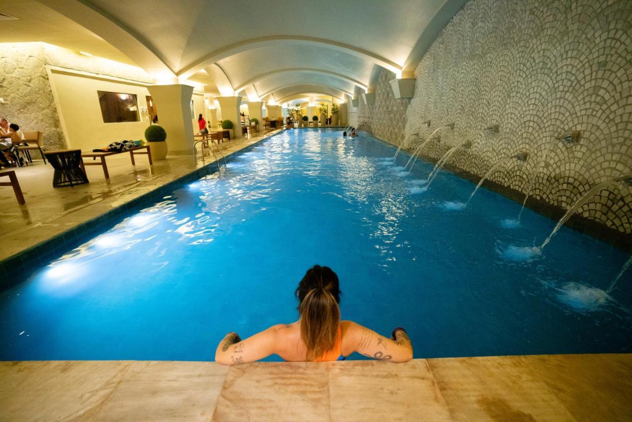 Buona Vitta Gramado Resort & Spa By Gramado Parks Exterior foto The photo shows an indoor swimming pool with a long, narrow design. The water is a clear blue, and there are features such as fountains along one side. In the foreground, a person with long hair is sitting on the edge of the pool, looking out over th